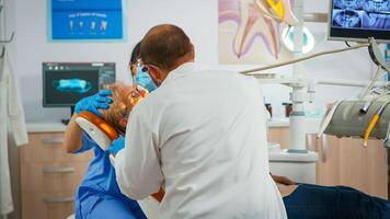 senior vrouw zittend in tandheelkundig kliniek nemen zorg van tand Gezondheid. orthodontist verlichting de lamp tot inspectie, geduldig opening mond aan het liegen Aan stomatologisch stoel terwijl verpleegster halpend dokter. foto