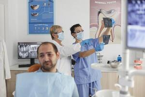 portret van geduldig met kiespijn aan het wachten voor medisch expertise gedurende stomatologisch examen in tandheelkunde kantoor kamer. in achtergrond medisch team analyseren tanden radiografie. geneeskunde onderhoud foto