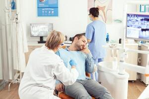 stomatoloog senior vrouw onderzoeken geduldig tand pijn gedurende stomatologie overleg. ziek Mens zittend Aan tandheelkundig stoel voorbereidingen treffen voor tandheelkunde chirurgie aan het wachten voor kiespijn behandeling foto