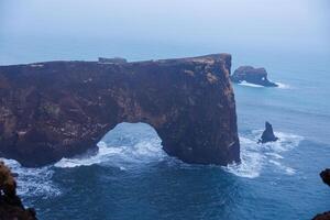 steen kustlijn boog Aan dyrholaey schiereiland met nordic mistig landschap en rotsachtig kliffen. spectaculair kustlijn steenachtig poort in IJsland, mooi wildernis met haven baai visie. foto