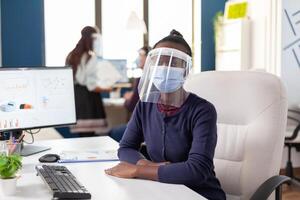 Afrikaanse zakenvrouw vervelend gezicht masker en vizier tegen covid19 op zoek Bij camera. multi-etnisch bedrijf team werken in financieel bedrijf met inachtneming van sociaal afstand gedurende globaal pandemie. foto