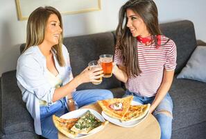 jong Dames hebben een lunch samen roosteren bieren en aan het eten pizza - gelukkig vrienden genieten van een avondeten zittend Aan bankstel in de leven kamer Bij huis - mensen, vriendschap, drankje, levensstijl concept foto