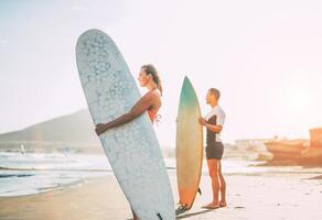 jong paar van surfers staand Aan de strand met surfplanken voorbereidingen treffen naar surfen Aan hoog golven gedurende een prachtig zonsondergang - mensen, levensstijl, sport concept - focus Aan vrouw foto