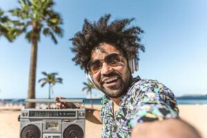 jong Latijns Mens hebben pret nemen selfie met mobiel smartphone terwijl luisteren muziek- met hoofdtelefoons en boombox Aan de strand gedurende zomer vakanties foto