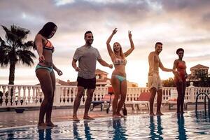 groep van gelukkig vrienden dansen Bij zwembad partij Bij zonsondergang - jong millennial mensen hebben pret in een tropisch luxe toevlucht - vakantie, zomer vakantie en jeugd levensstijl concept foto