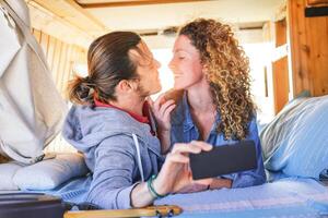 gelukkig paar hebben een romantisch datum in wijnoogst busje met hout interieur - Mens en vrouw nemen selfie met mobiel telefoon terwijl zoenen gedurende hun reis - reis, liefde, technologie concept foto