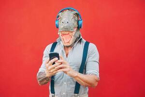 senior mode Mens vervelend t-rex masker gebruik makend van mobiel smartphone luisteren muziek- met hoofdtelefoons - gek bizar hipster vent hebben pret met nieuw technologie - grappig en absurd surrealistische concepten foto