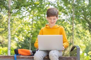 glimlachen tiener jongen werken Aan laptop. Holding en gebruik makend van een laptop voor netwerken Aan een zonnig voorjaar dag, buitenshuis. foto