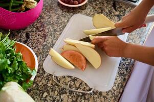 bijgesneden visie van vrouw handen snijden aardappel Aan een snijdend bord terwijl voorbereidingen treffen avondeten in de huis keuken foto