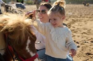 jong vrouw moeder en haar aanbiddelijk weinig kind meisje, een lief dochter glimlachen en strelen de pony foto