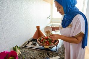 Marokkaans vrouw huisvrouw in blauw hijab en authentiek jurk, Koken vers groenten in tajine klei pot in de keuken foto