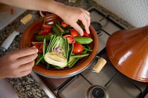 visie van bovenstaand van huisvrouw handen zetten ingrediënten terwijl Koken tajine met heerlijk biologisch groenten foto