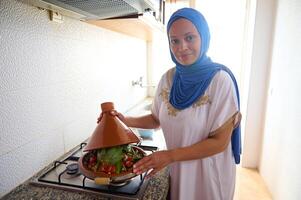 glimlachen midden- oostelijk huisvrouw met hoofd gedekt in hijaab, staand door fornuis, Koken Marokkaans tajine met groenten foto