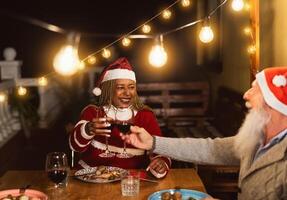 gelukkig multiraciaal senior vrienden roosteren met rood wijn bril gedurende Kerstmis vakantie avondeten viering Aan patio huis partij foto