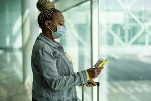 Afrikaanse senior vrouw in luchthaven terminal met bagage vervelend gezicht masker gedurende coronavirus pandemisch foto