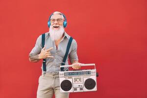 senior gek Mens luisteren muziek- met hoofdtelefoons en wijnoogst boombox buitenshuis - hipster mannetje hebben pret leven in Verleden tijd - ouderen mensen levensstijl werkzaamheid - rood achtergrond foto