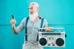 gelukkig senior Mens gebruik makend van mobiel telefoon terwijl Holding wijnoogst boombox buitenshuis - mode hipster mannetje hebben pret luisteren muziek- en gebruik makend van smartphone buiten - technologie en ouderen levensstijl concept foto