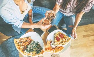 jong Dames juichen bier en aan het eten pizza in hun appartement - twee meisjes hebben een lunch samen met eigengemaakt voedsel - concept van mensen, vrienden en levensstijl foto