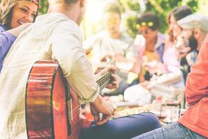 groep van vrienden hebben een picknick in een park buitenshuis - gelukkig jong vrienden genieten van picknick spelen gitaar, het zingen en drinken wijn aan het eten voedsel - recreatie concept - focus Aan Mens arm met gitaar foto