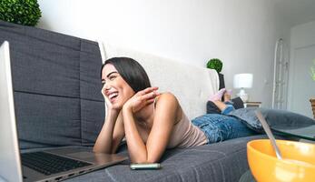 jong vrouw aan het liegen Aan sofa aan het kijken Aan computer - gelukkig meisje hebben pret gebruik makend van laptop Bij huis - jeugd millennial generatie levensstijl en technologie concept foto