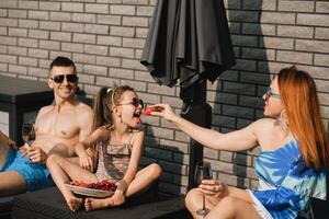 een gelukkig familie in zwemkleding zonnebaadt Aan hun terras in zomer. mam feeds haar dochter aardbeien foto