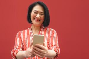 gelukkig Aziatisch vrouw gebruik makend van mobiel smartphone buitenshuis - vrouw senior hebben pret met nieuw modieus technologie sociaal media apps - ouderen levensstijl en tech oud mensen verslaafd concept - rood achtergrond foto