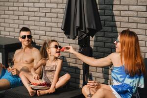 een gelukkig familie in zwemkleding zonnebaadt Aan hun terras in zomer. mam feeds haar dochter aardbeien foto