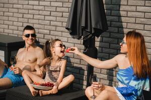 een gelukkig familie in zwemkleding zonnebaadt Aan hun terras in zomer. mam feeds haar dochter aardbeien foto