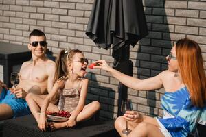 een gelukkig familie in zwemkleding zonnebaadt Aan hun terras in zomer. mam feeds haar dochter aardbeien foto