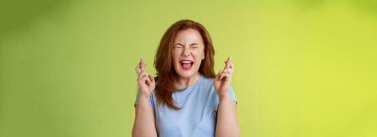 vrouw wil winnen slecht. enthousiast Lucky roodharige middelbare leeftijd Jaren 50 vrouw pleiten smeken god maken droom komen waar kruis vingers mooi zo geluk wensen Gesloten ogen Open mond opwinding groen achtergrond foto