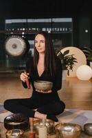 een vrouw in de lotus positie gebruik makend van een het zingen kom binnenshuis . ontspanning en meditatie. geluid therapie, alternatief geneesmiddel. boeddhistisch genezing praktijken foto