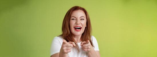 u deed het beste. vriendelijk blij enthousiast roodharige gember middelbare leeftijd vrouw richten inhoudsopgave vingers camera vinger pistool gebaar glimlachen breed feliciteren juichen collega staan groen achtergrond foto