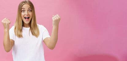 opgewonden vrolijk en optimistisch charmant vrouw met eerlijk haar- in wit t-shirt verhogen vuisten in zege en triomf schreeuwen Ja in succes en verbazing staand ondersteunend tegen roze achtergrond foto