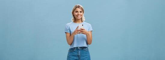 nieuw telefoon is fantastisch. portret van tevreden gelukkig jong modern blond vrouw in modieus kleding met blond kort kapsel glimlachen vrolijk van genot Holding smartphone in de buurt borst sms'en of gebruik makend van apps foto