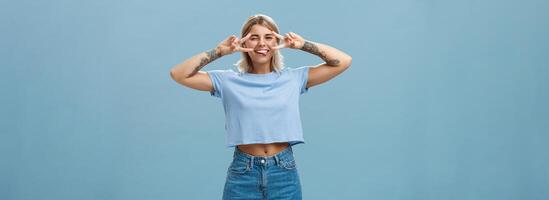 genieten leven en droom. portret van blij mooi zorgeloos Europese vrouw leerling met blond kort kapsel knipogen plakken uit tong vermaakt en tonen vrede tekens over- ogen in de buurt blauw muur foto