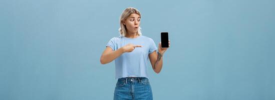 u zou moeten downloaden app. portret van versteld staan geschokt aantrekkelijk blond meisje in modieus zomer t-shirt Holding smartphone hijgen van verrassing richten Bij apparaatje scherm poseren over- blauw achtergrond foto