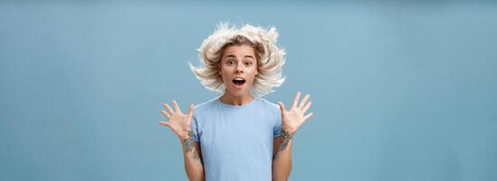 van je middel omhoog schot van onder de indruk verrast aantrekkelijk en schattig meisje met blond haar- drijvend in lucht en getatoeëerd arm jumping met verspreiding verheven palmen en geopend mond over- blauw achtergrond foto