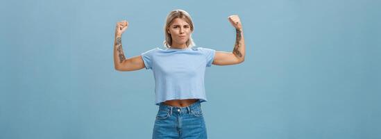 sterk en krachtig mooi sport vrouwen met tatoeages in t-shirt en shorts verhogen armen tonen groot spieren en biceps glimlachen trots terwijl opscheppen met fysiek sterkte over- blauw muur foto