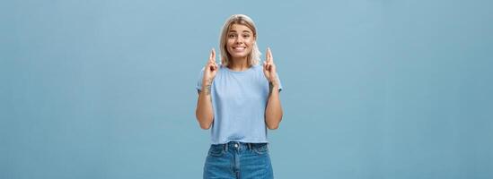 hoop droom komen waar spoedig. portret van emotioneel gelukkig en optimistisch aantrekkelijk jong Europese vrouw met blond haar- en gebruind huid kruispunt vingers voor mooi zo geluk en glimlachen over- blauw muur foto