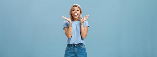 opgewonden enthousiast gelukkig vrouw in modieus t-shirt en shorts klappen handen van verbazing glimlachen breed en staren met bewondering Bij camera wezen verrast en blij over- blauw muur foto