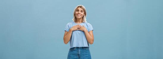 zo zoet bedankt. aangeraakt mooi en emotioneel blond meisje in t-shirt en shorts met gebruind huid Holding palmen Aan borst, zuchtend en glimlachen blij wezen dankbaar en dankbaar over- blauw muur foto