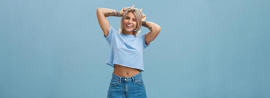 studio schot van schattig aantrekkelijk en blij elegant blond vrouw met tatoeages Aan armen knipogen en glimlachen breed Holding inhoudsopgave vingers Aan hoofd maken hoorns staand in zomer kleding over- blauw muur foto