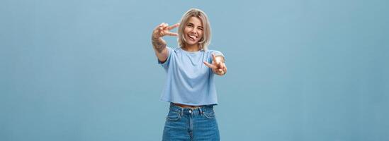 mooi getatoeëerd meisje genieten van weekends plakken uit tong blij knipogen Bij camera en glimlachen tonen vrede of zege gebaar met getrokken armen gevoel gelukkig poseren over- blauw achtergrond foto