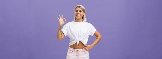 uitstekend ik Leuk vinden het. portret van tevreden mooi gelukkig meisje met eerlijk haar- in wit t-shirt en shorts tonen OK of perfect gebaar en glimlachen breed Holding hand- Aan taille over- Purper muur foto