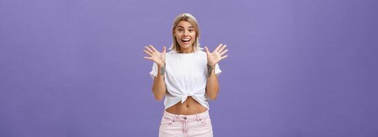 binnen- schot van verheugd en geamuseerd aantrekkelijk elegant stedelijk vrouw met blond haar- in elegant wit t-shirt en zomer shorts verhogen palmen van verrassing en verbazing staand over- Purper achtergrond foto