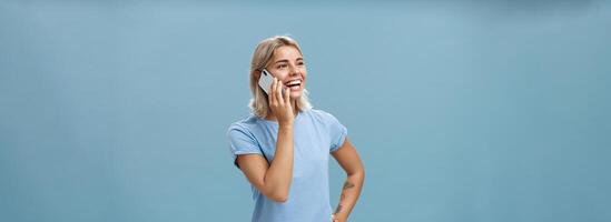 van je middel omhoog schot van sociaal geamuseerd en gelukkig aantrekkelijk Kaukasisch blond haar vrouw in gewoontjes t-shirt staand half omgedraaid staren links met hand- Aan heup terwijl pratend Aan smartphone over- blauw achtergrond foto