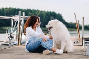 een gelukkig vrouw met een groot wit hond zit Aan een pier door de zee Bij zonsondergang foto