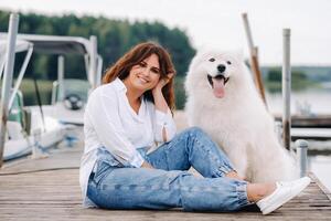 een gelukkig vrouw met een groot wit hond zit Aan een pier door de zee Bij zonsondergang foto