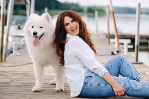 een gelukkig vrouw met een groot wit hond zit Aan een pier door de zee Bij zonsondergang foto