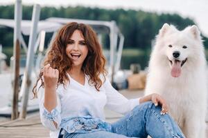 een gelukkig vrouw met een groot wit hond zit Aan een pier door de zee Bij zonsondergang foto